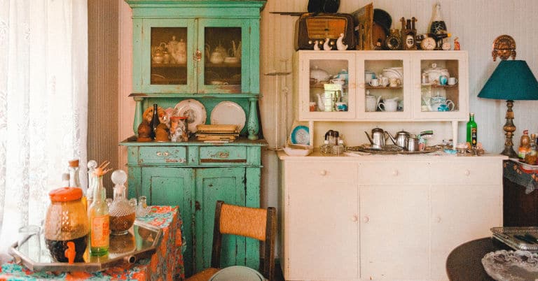 A kitchen with old furniture and items in and on cabinets and shelves.